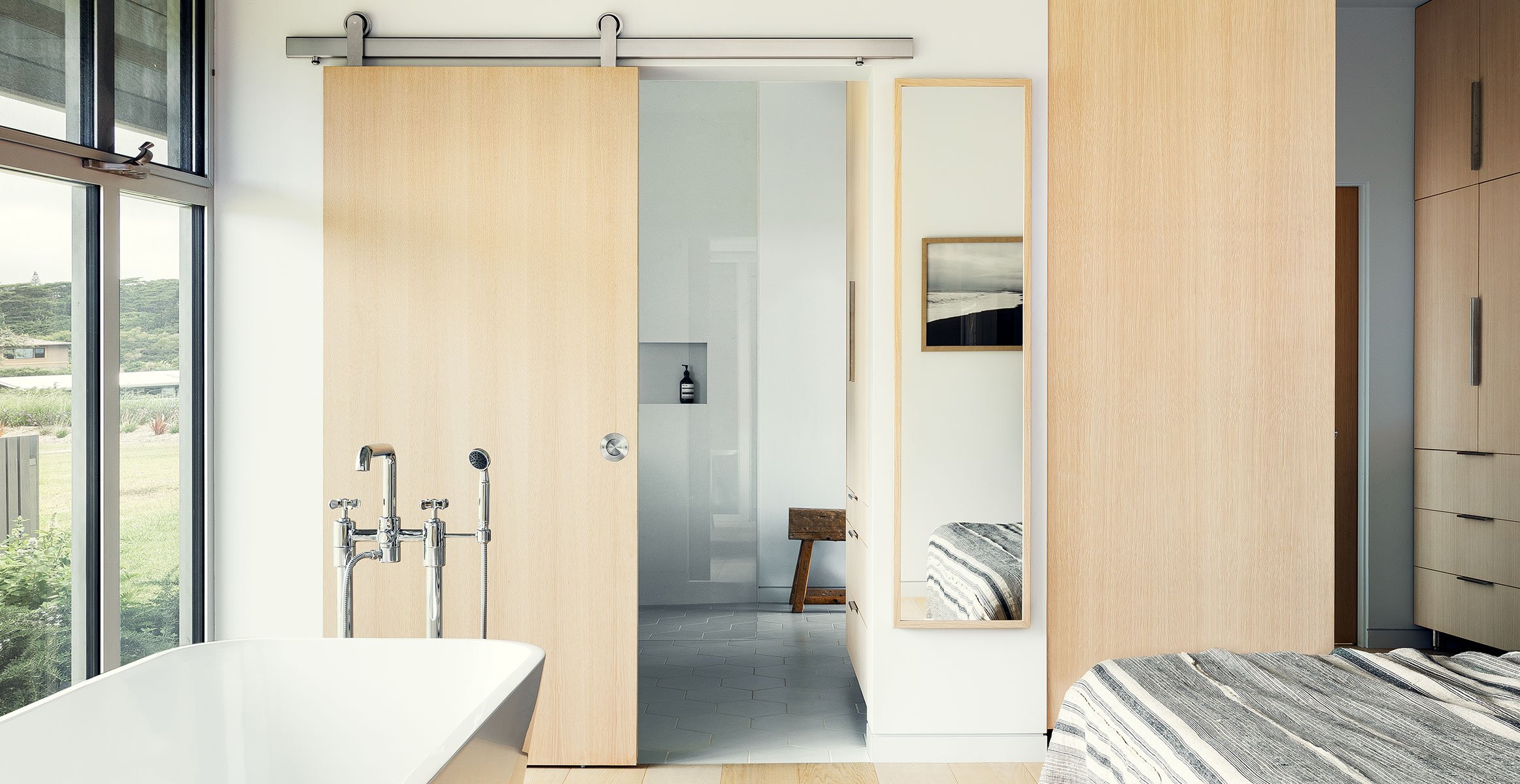 Modern sliding barn door hardware in brushed stainless finish by Krownlab on natural Ash door in bathroom with freestanding tub.